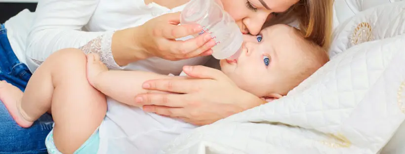 Mother bottle-feeding baby on bed.