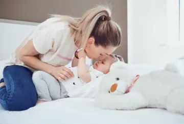 Mother kissing baby on bed with teddy.