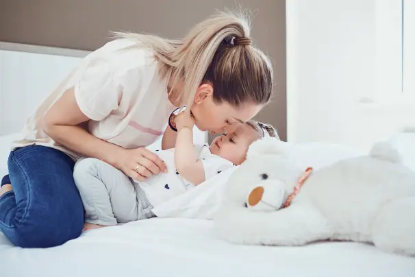 Mother kissing baby on bed with teddy.